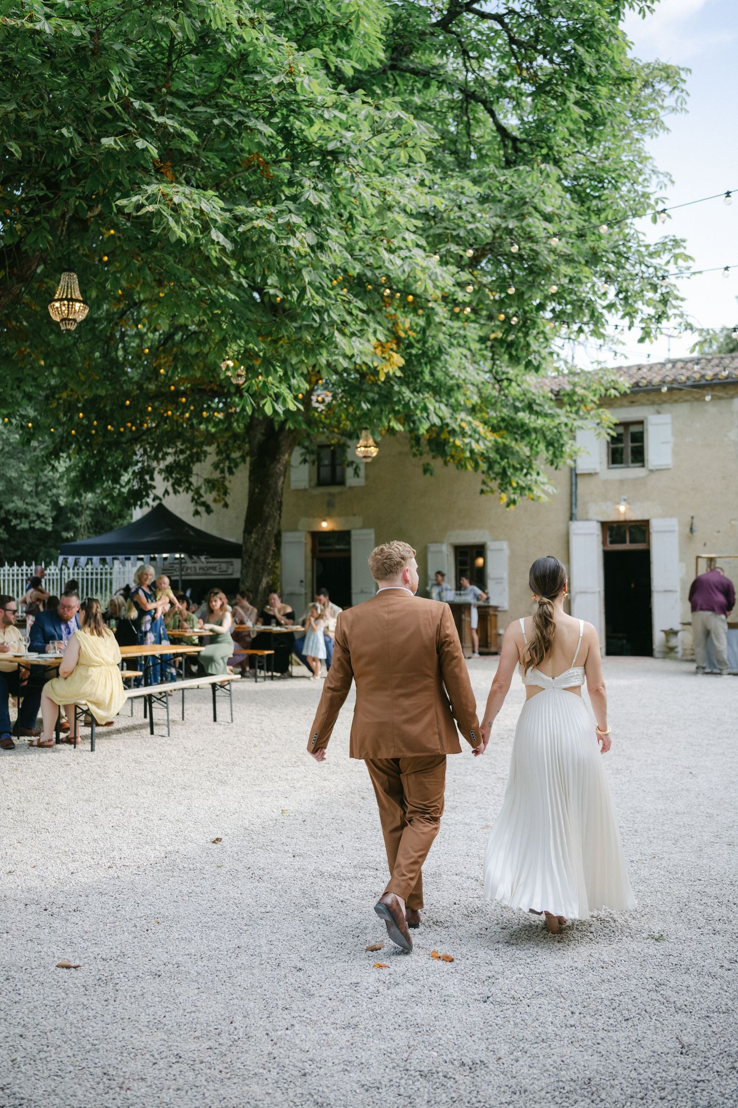 Bride and groom walking hand in hand through a rustic outdoor wedding venue with guests seated at long tables under a large tree.