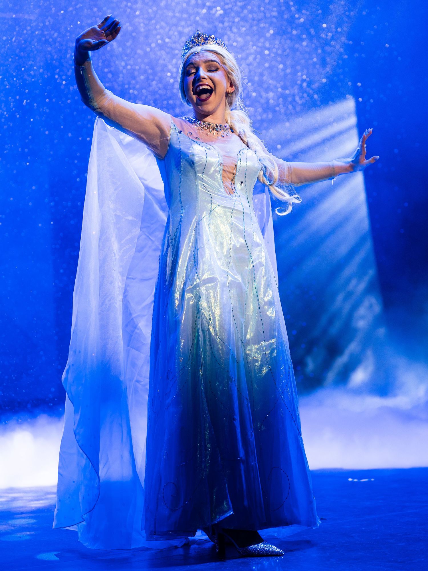 Performer in sparkling ice queen costume singing passionately on a blue-lit theatrical stage.
