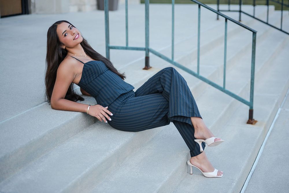 Woman in striped navy jumpsuit and white heels sitting gracefully on outdoor steps, smiling elegantly.