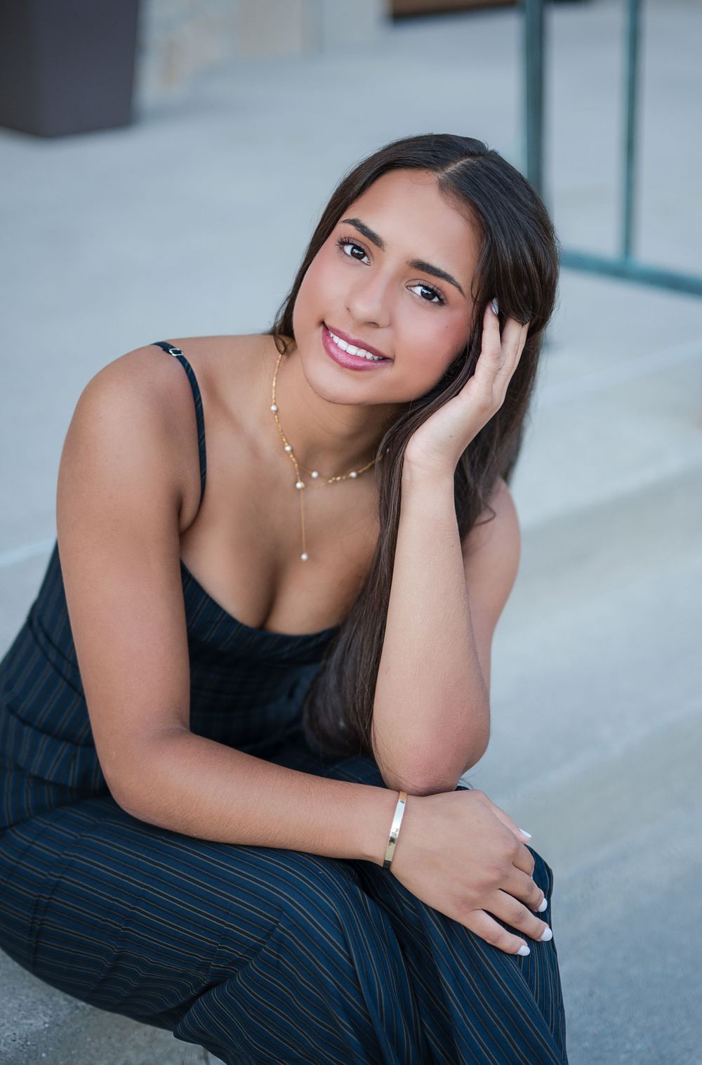 Young woman in navy pinstripe outfit sits on steps, smiling, wearing gold jewelry and a silver bracelet.