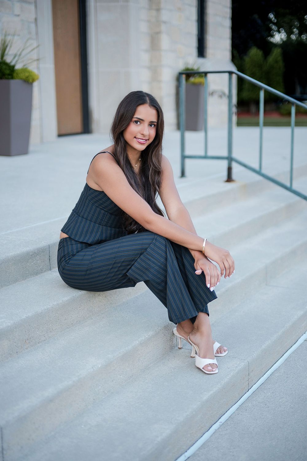 Young woman in pinstripe pants and black top sitting on outdoor steps, smiling, wearing white heels.