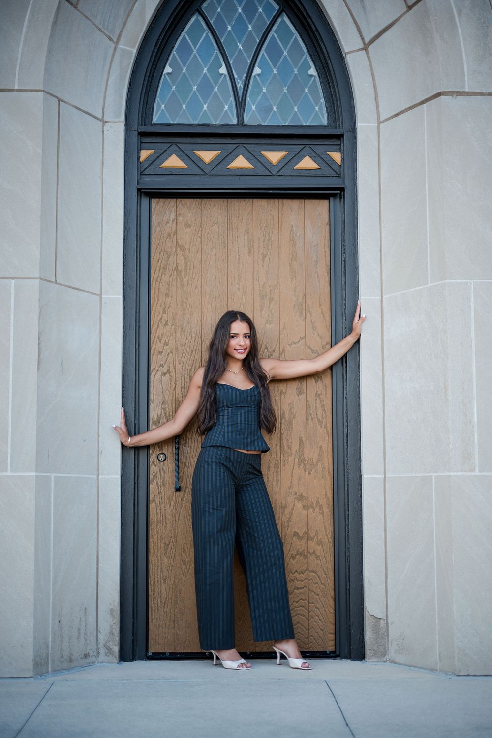 Young woman in striped pantsuit posing confidently in an arched wooden doorway of a stone building.