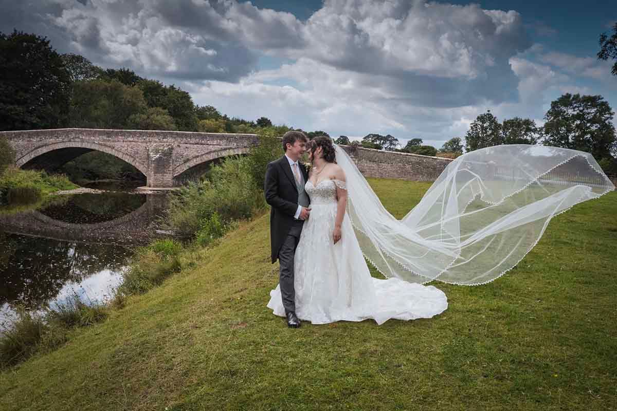Bride and groom embrace on green lawn with veil flowing in wind, beside a stone bridge reflected in calm water.