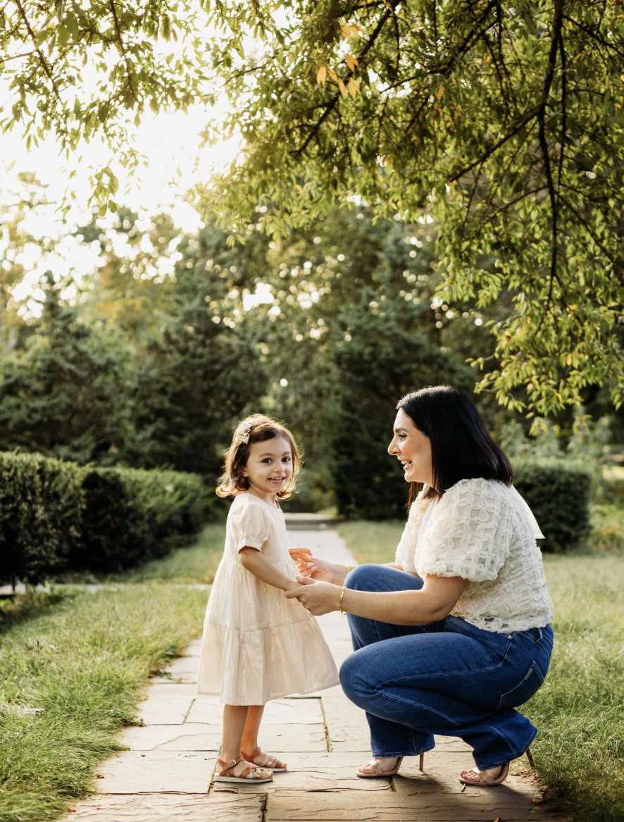A mother crouches on a garden path, smiling at her toddler daughter in a white dress on a sunny day.