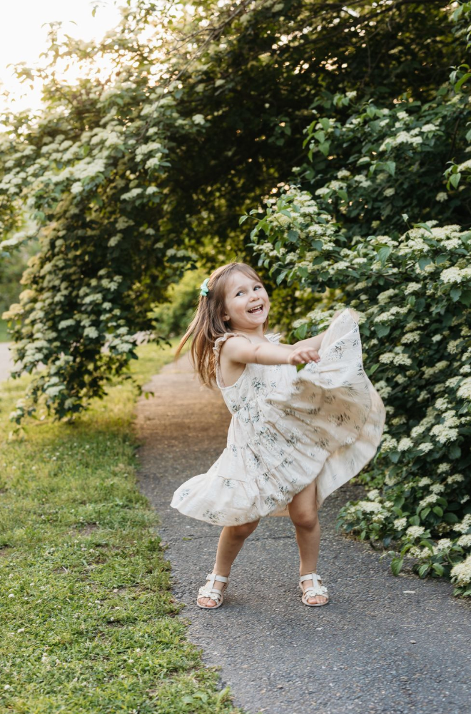 A joyful little girl in a floral dress twirls on a garden path surrounded by lush green trees.