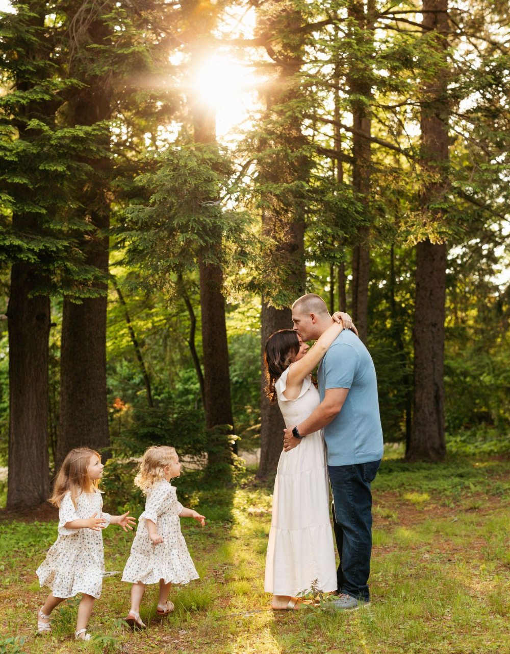 Family portrait of parents embracing while two young daughters dance together in a sunlit forest clearing.