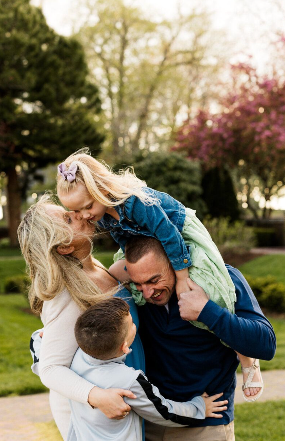 Happy family of four sharing a joyful moment outdoors, with parents hugging children in a lush green park setting.