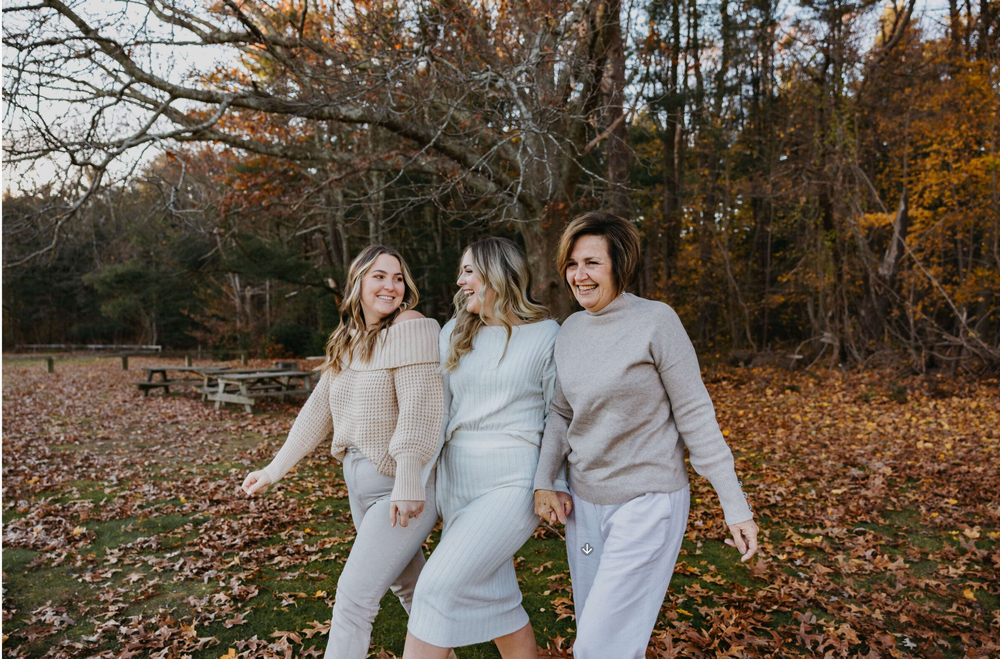 Three women in neutral tones laugh and walk together through a fall park covered in colorful autumn leaves.