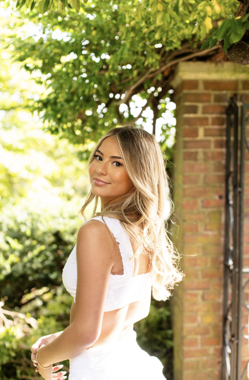 Young woman with long blonde hair wearing a white dress poses outdoors near a brick wall with lush green foliage.
