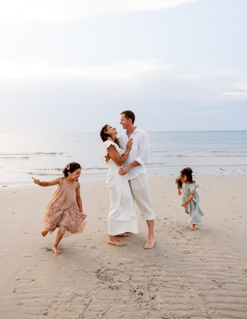 A joyful family dancing on a sandy beach, with two young girls spinning around their parents near the ocean.