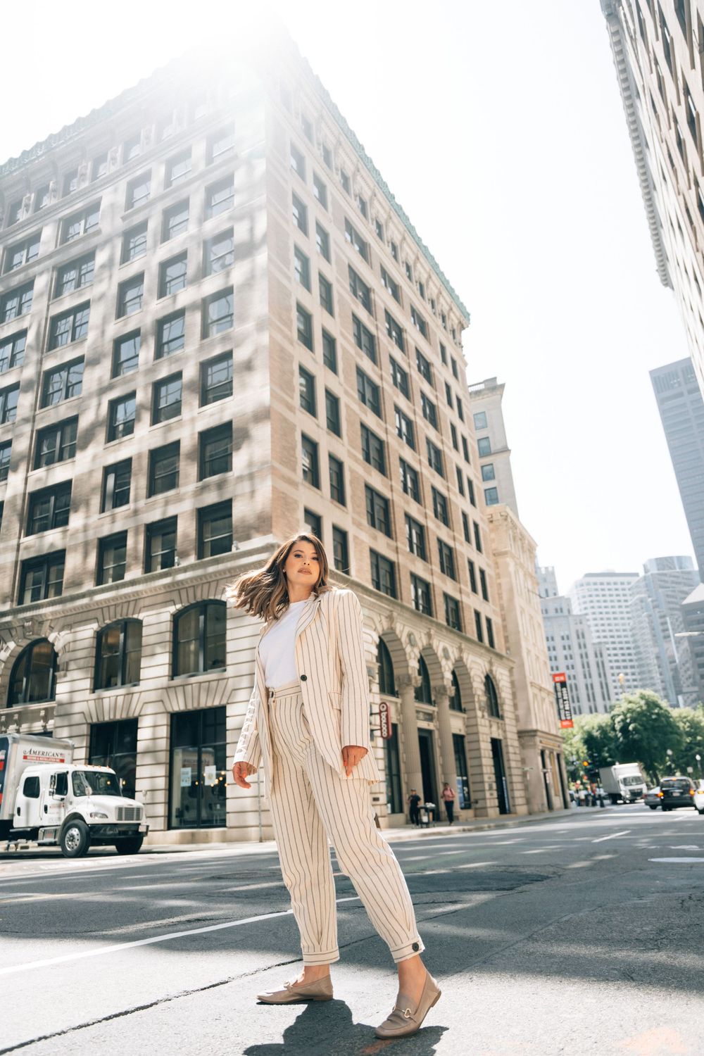 Woman in striped suit walking confidently on a sunny urban street with tall city buildings in the background.