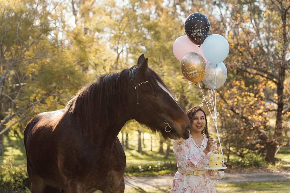 Woman in floral dress holding colorful balloons stands beside a brown horse in Sydney Hawkesbury during a photoshoot with a horse