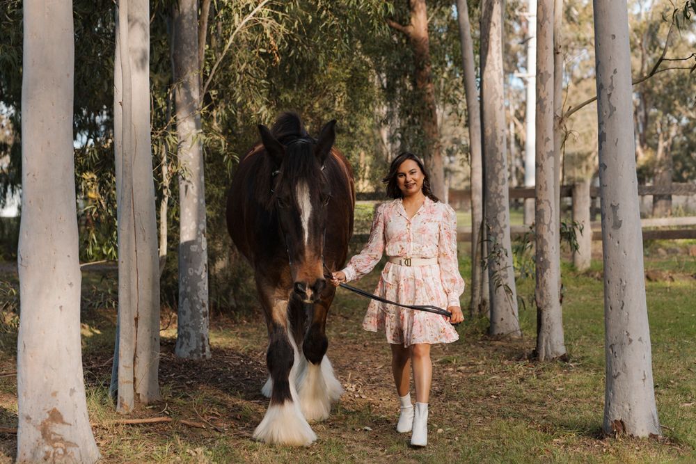Woman in floral dress leading a large Shire horse through a tree-lined path in Sydney during a photoshoot with a horse
