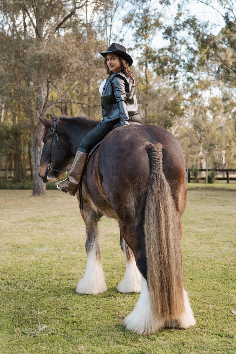 A smiling woman in a black cowboy hat and leather jacket rides a large brown Shire horse on a green pasture in Sydney during a photoshoot