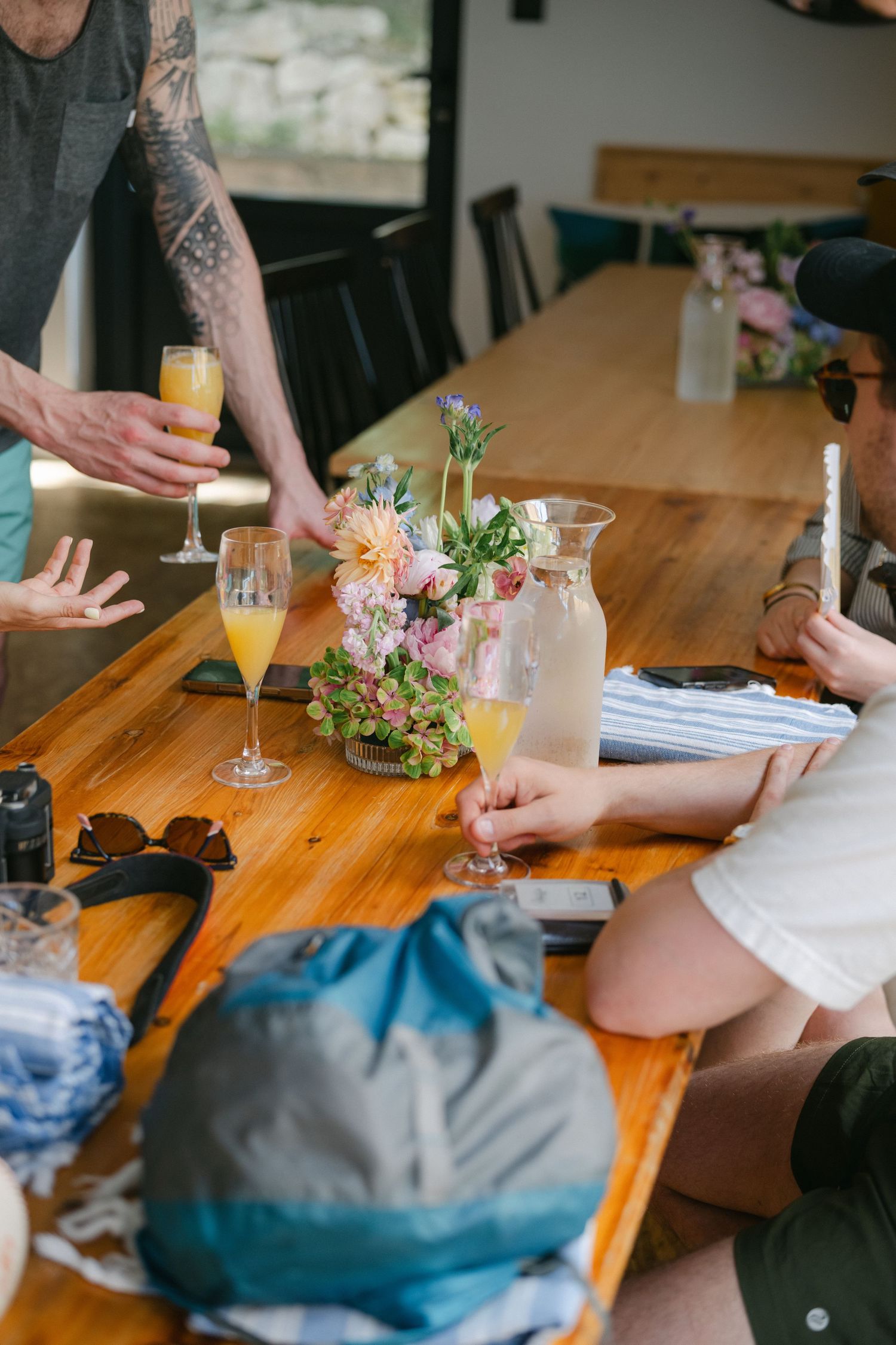People gathered around a rustic wooden table with mimosas, fresh flowers, and a lemonade pitcher during a sunny brunch gathering.