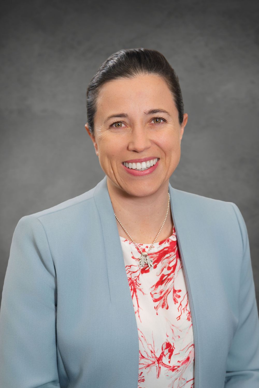 Professional headshot of a smiling woman in a light blue blazer and floral blouse against a gray background. Quad Cities Photography