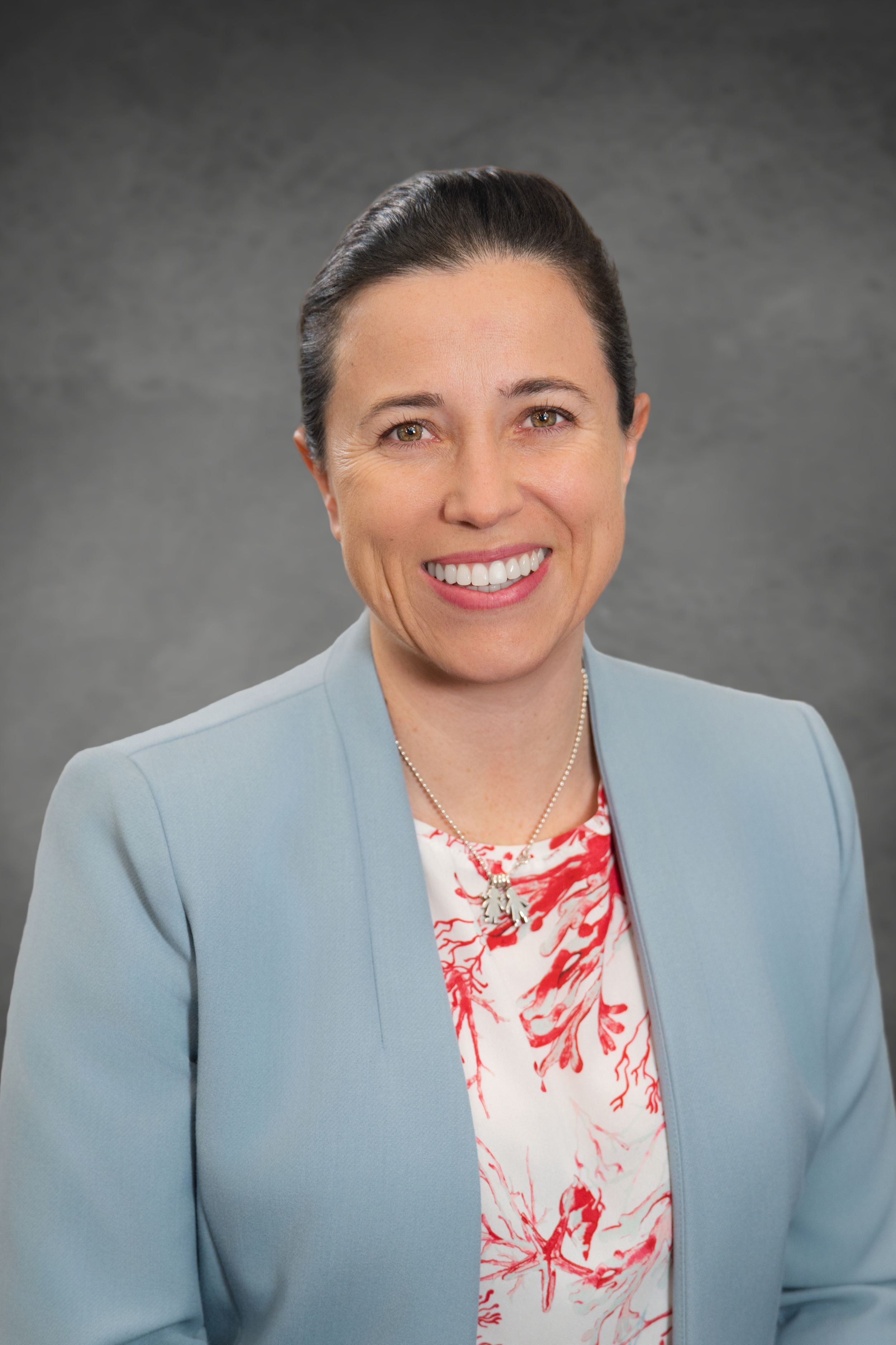 Professional headshot of a smiling woman in a light blue blazer and floral blouse against a gray background. Quad Cities Photography