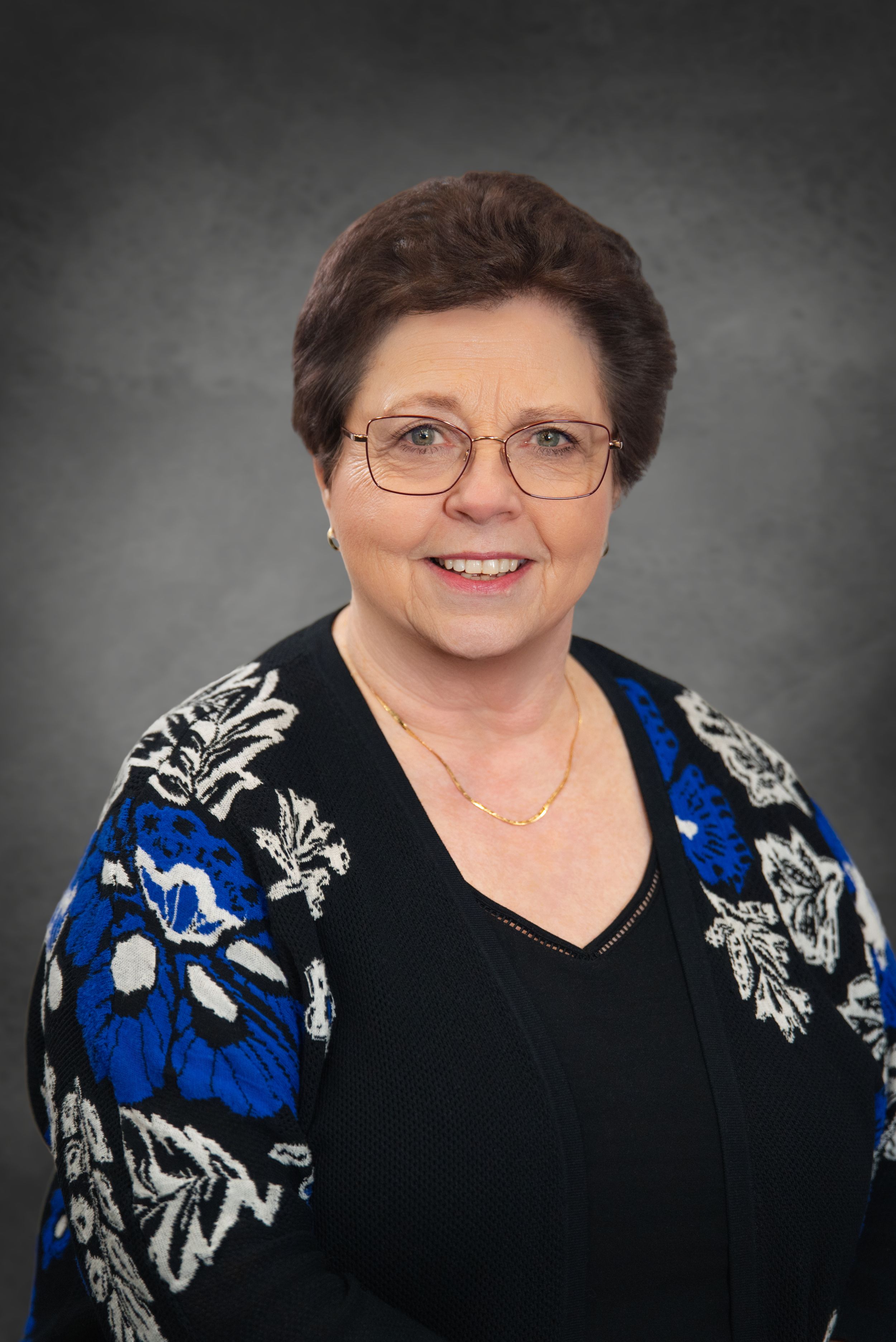 Smiling older woman with glasses wearing a floral blue and black cardigan, professional headshot on gray background.  Quad Cities Photograph
