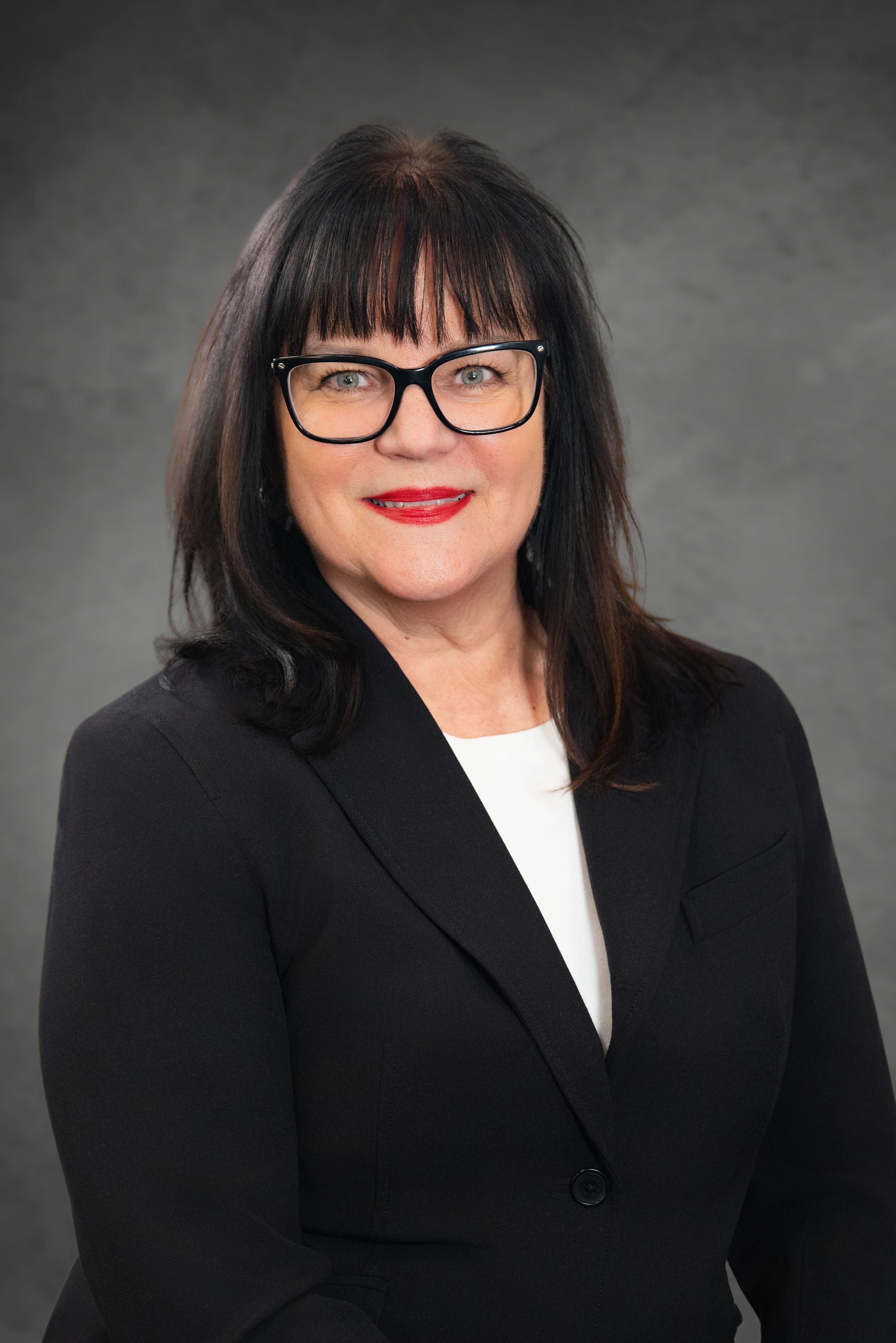 Professional headshot of a woman with dark hair, red lips, and black-rimmed glasses wearing a black blazer. Quad Cities Photography