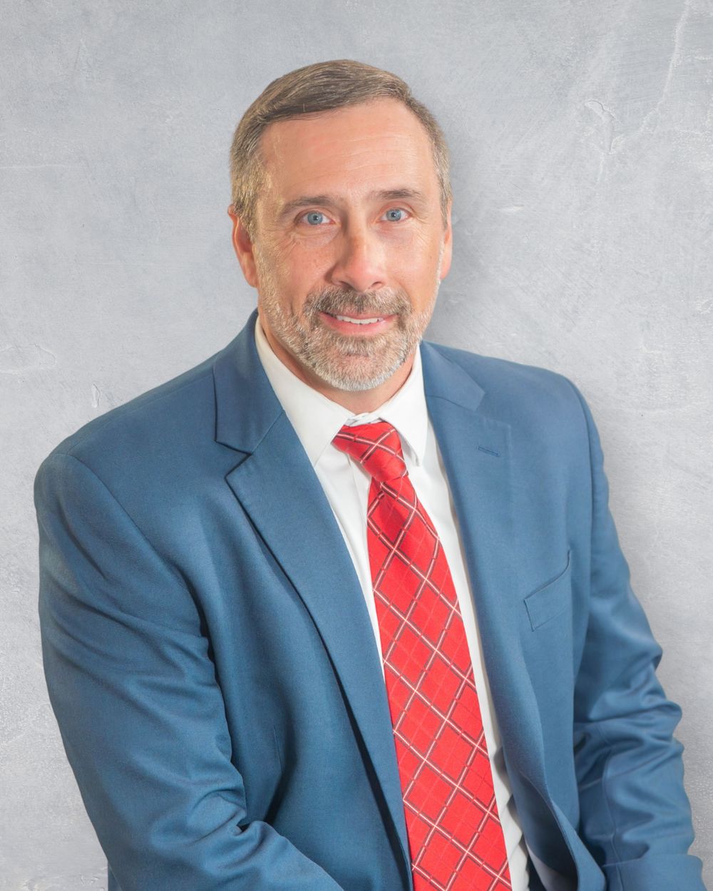 Professional headshot of a middle-aged man with gray beard wearing a blue suit and red tie against gray background. Quad Cities Photography