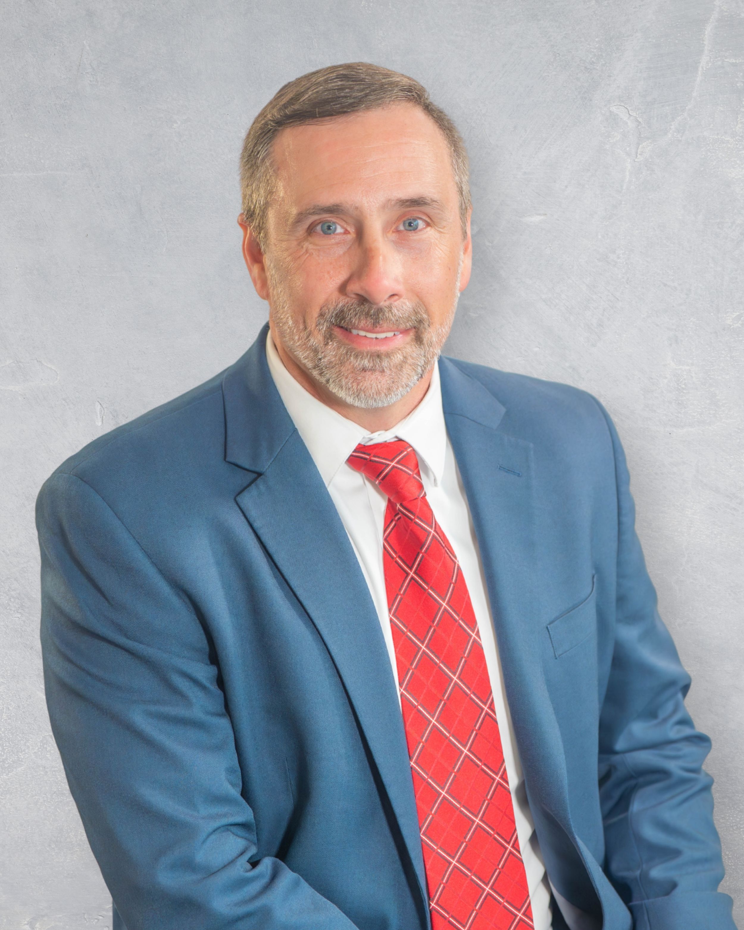 Professional headshot of a middle-aged man with gray beard wearing a blue suit and red tie against gray background. Quad Cities Photography