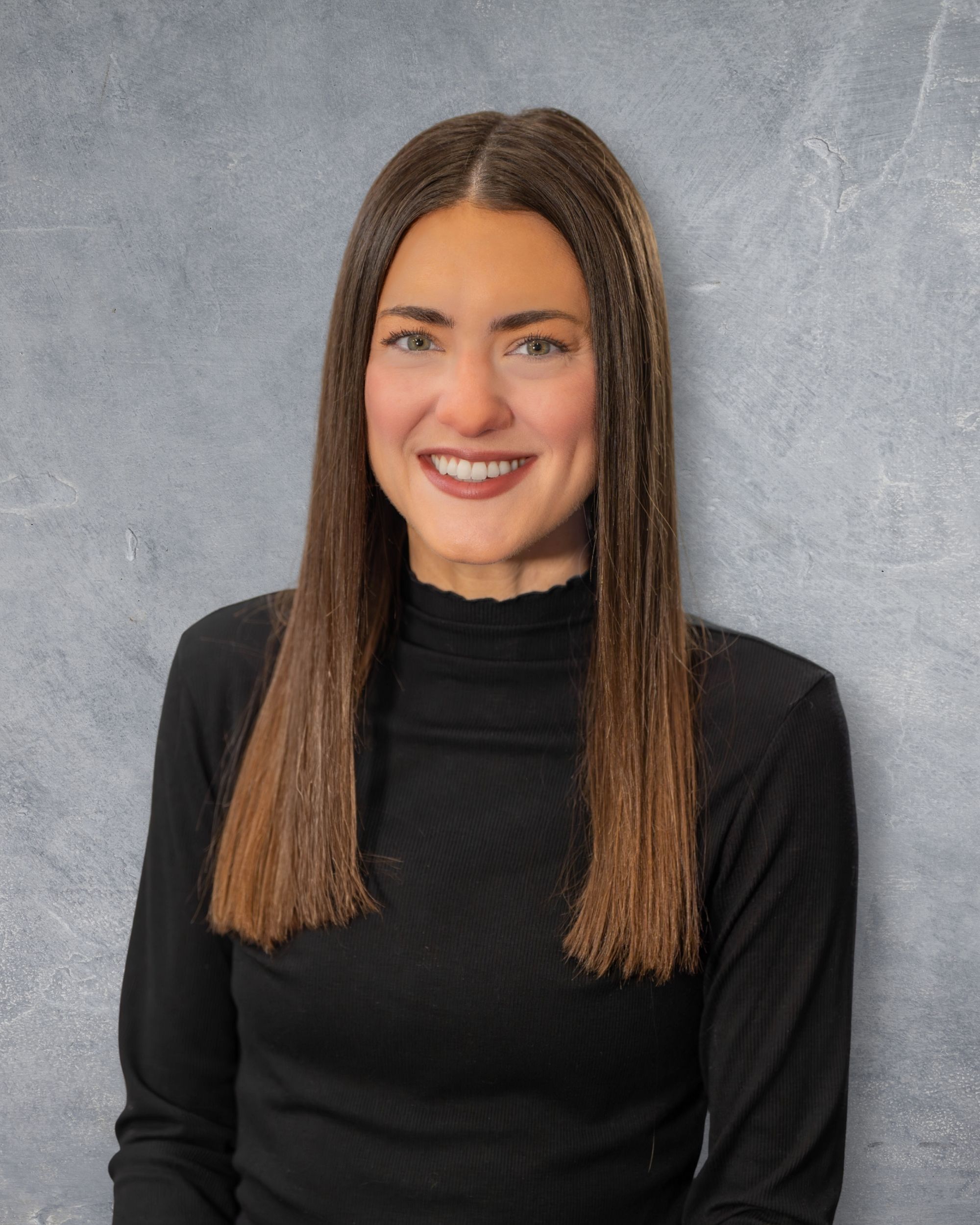 Professional headshot of a smiling woman with long brown hair wearing a black turtleneck top against a gray background.  Quad Cities Photo