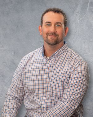 Professional headshot of a smiling man with a beard wearing a plaid shirt against a gray background.  Quad Cities Photography