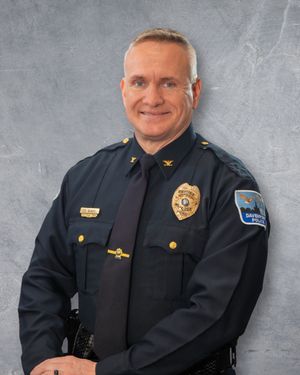 Police officer in dark navy uniform with gold badge and rank insignia smiling professionally against gray background.  Quad Cities Photo