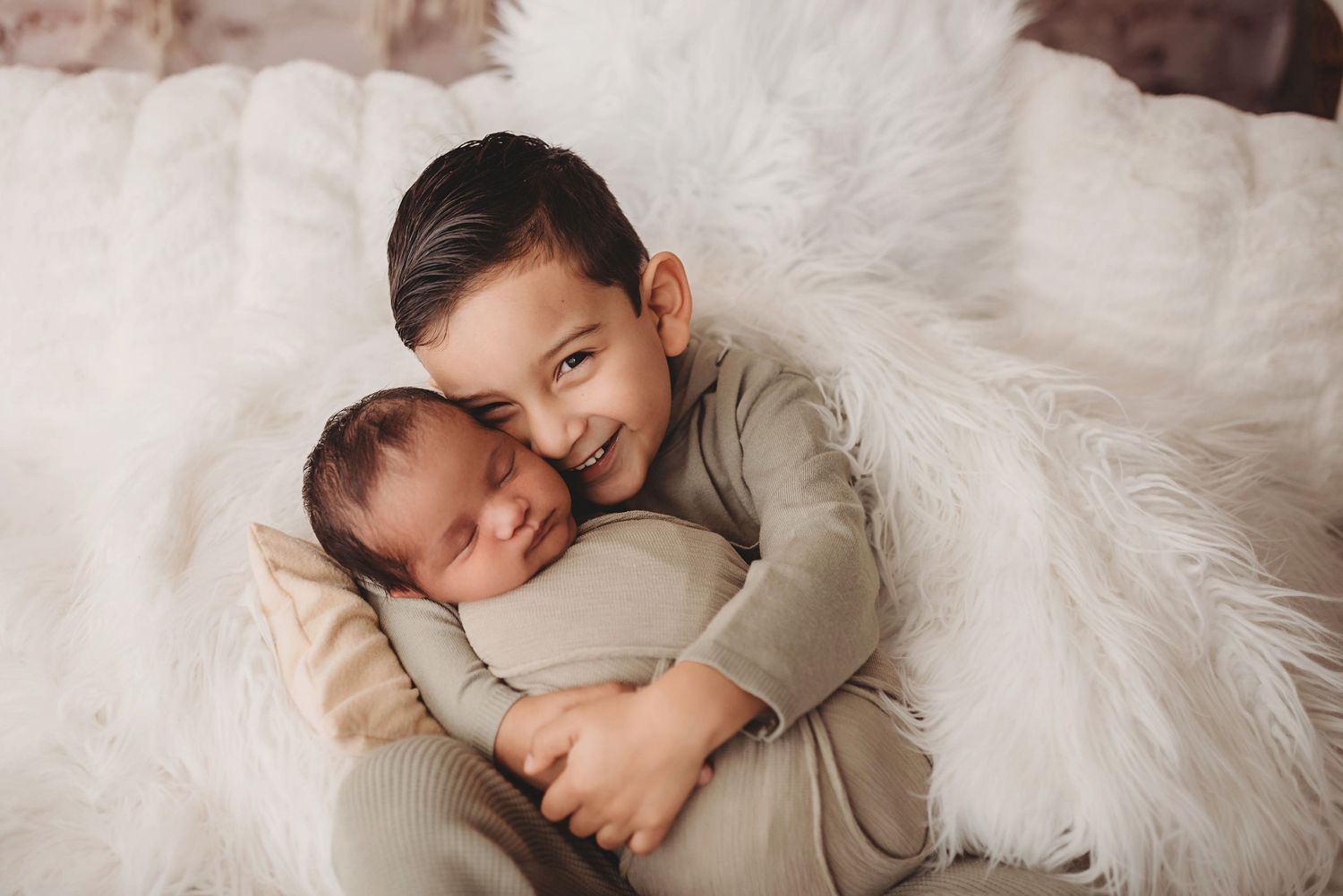 Young boy smiling while gently hugging a sleeping newborn baby on a white fluffy blanket.