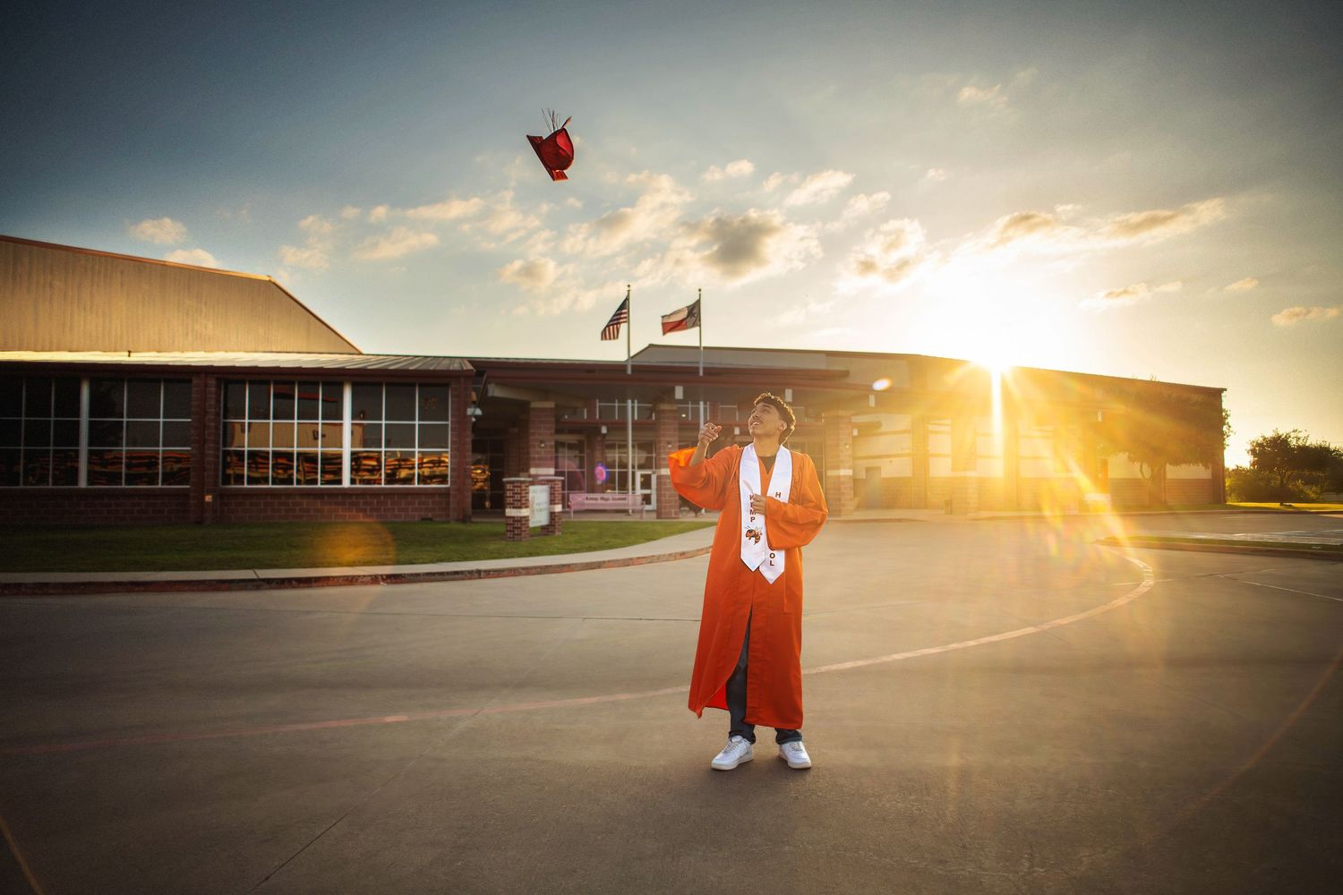 Graduate in orange cap and gown tosses cap in air outside school building at golden sunset.