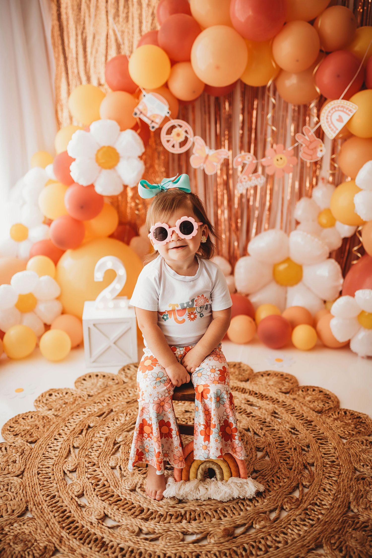 Toddler girl in retro floral bell bottoms and round sunglasses poses at a groovy 2nd birthday party with balloon arch.