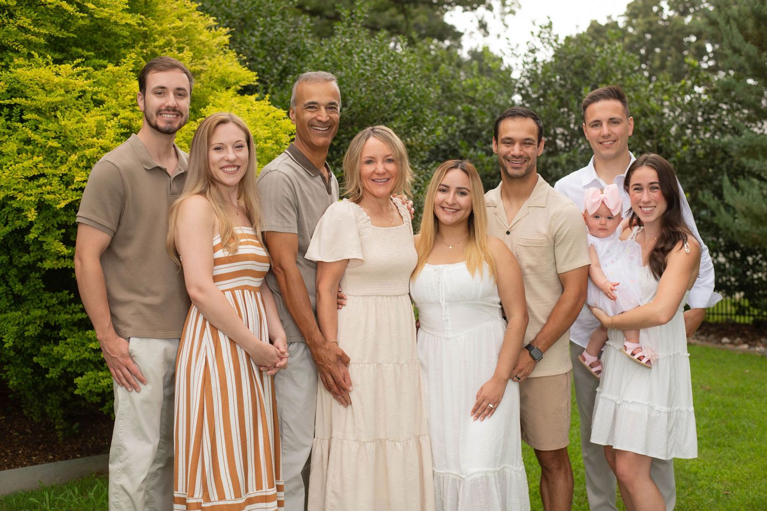 A joyful family of eight poses outdoors in a lush green garden, dressed in elegant summer attire.