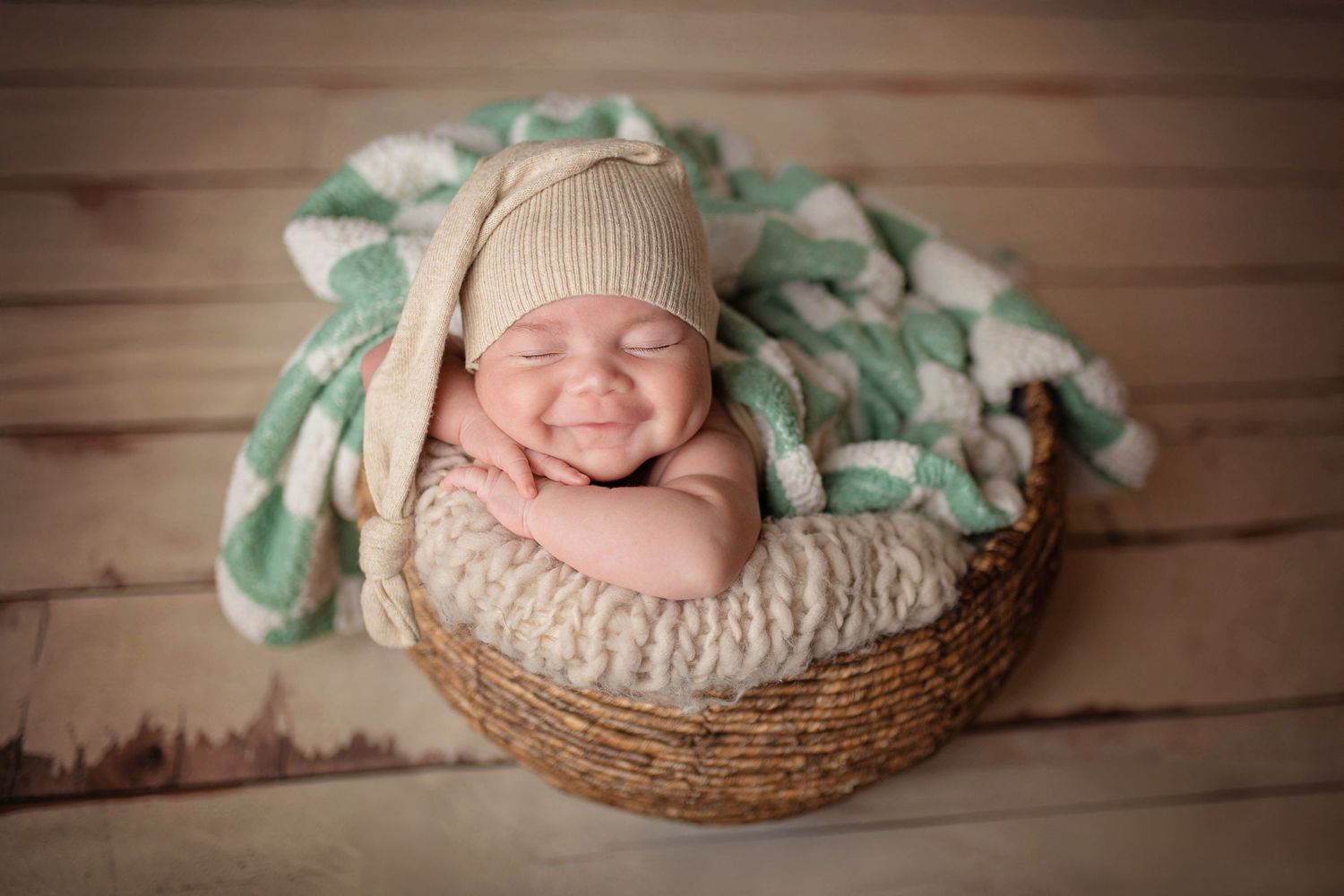 Smiling newborn baby in a wicker basket wearing a beige knit hat, nestled in a mint green chunky knit blanket.