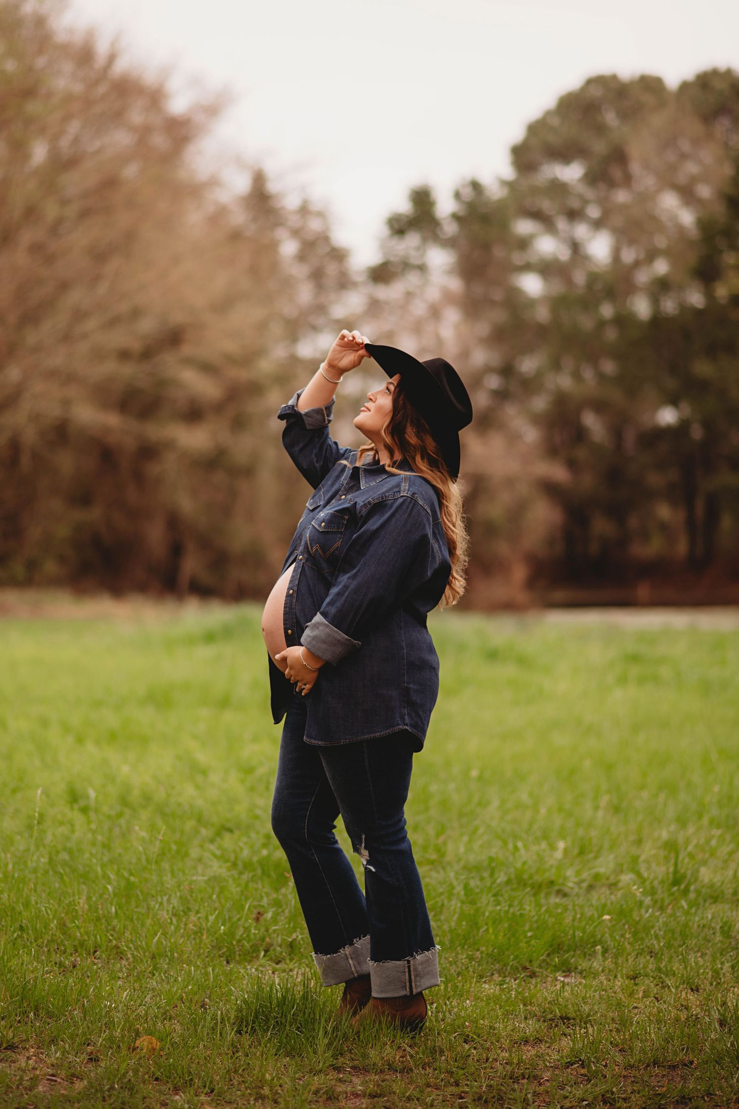 Pregnant woman in denim shirt and black cowboy hat posing in green field with trees in background.