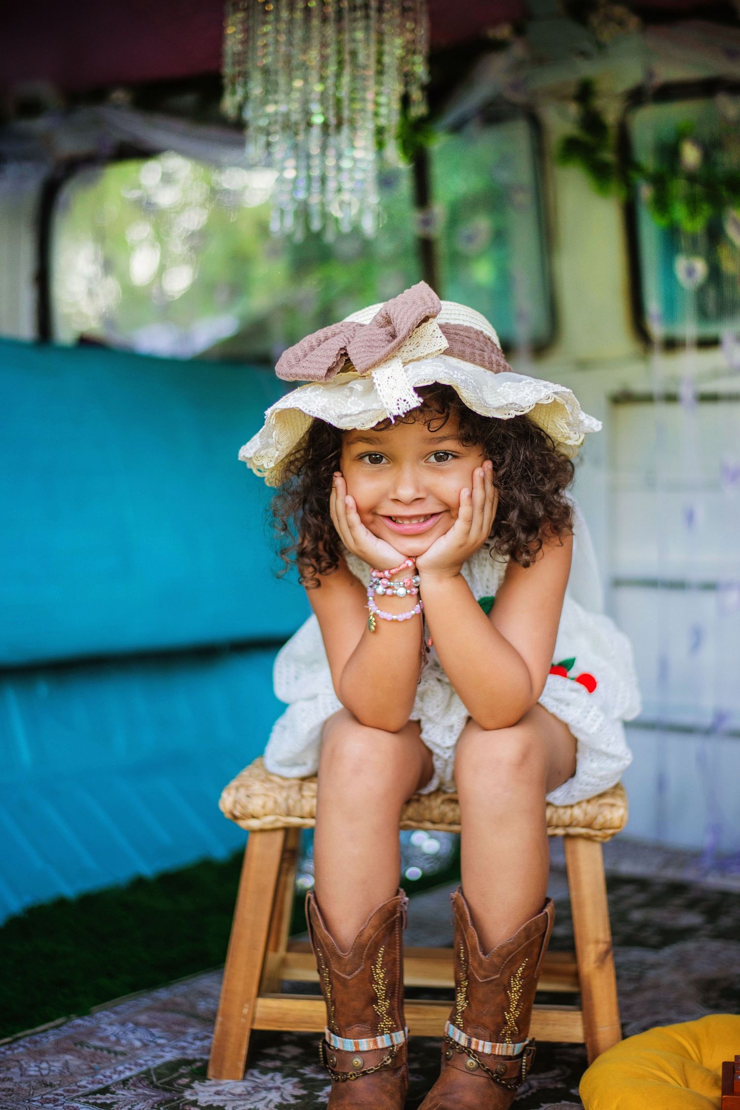 A smiling young girl in a wide-brim hat and cowboy boots sits on a stool with hands on cheeks.