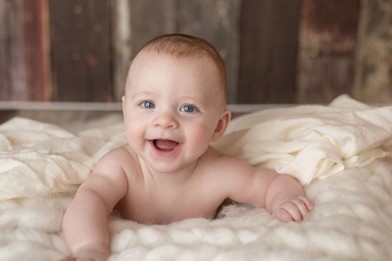 Smiling baby lying on a white knit blanket, blue eyes, happy expression, infant portrait photography.