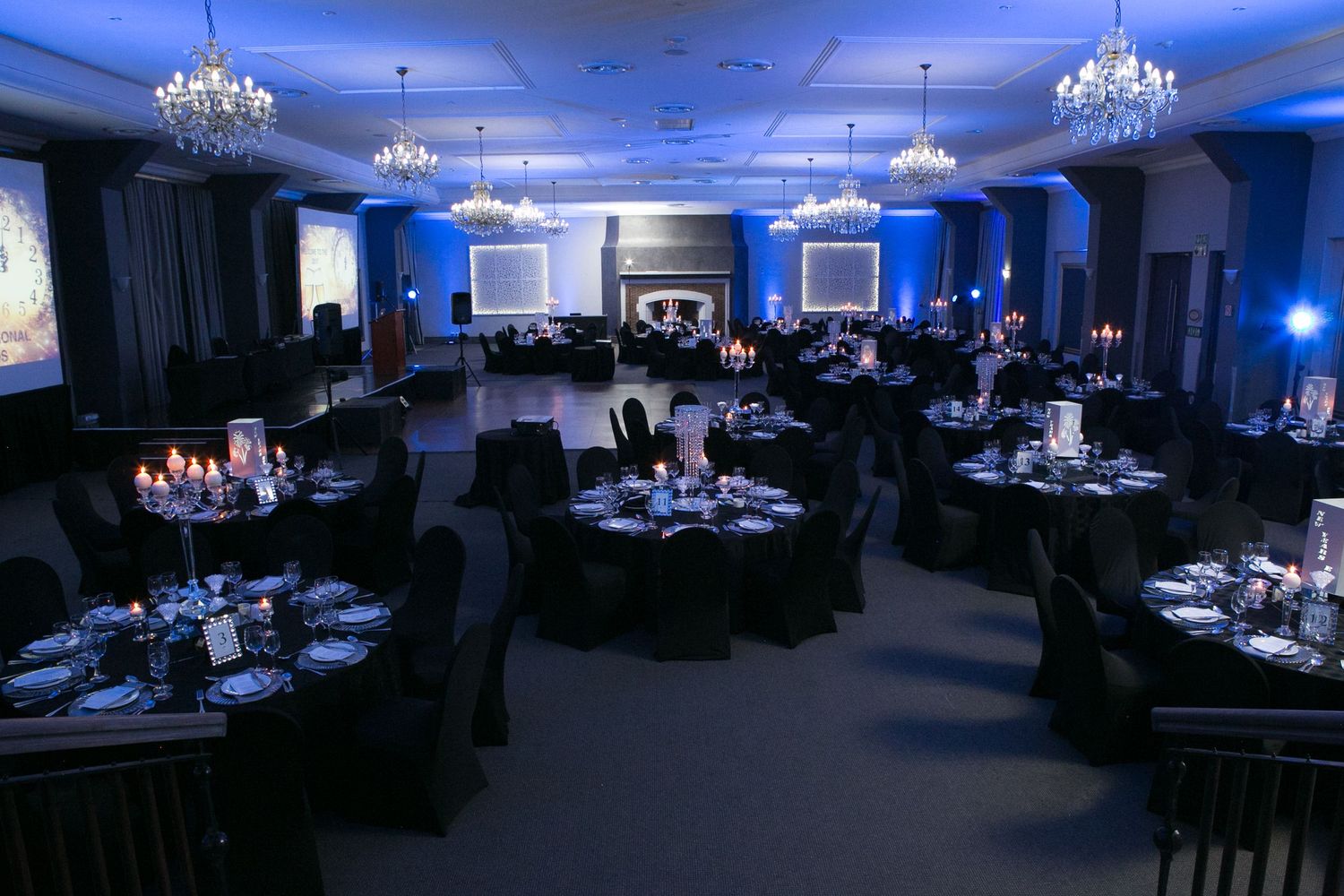 Elegant banquet hall with black round tables, crystal chandeliers, and blue ambient lighting for a formal event.