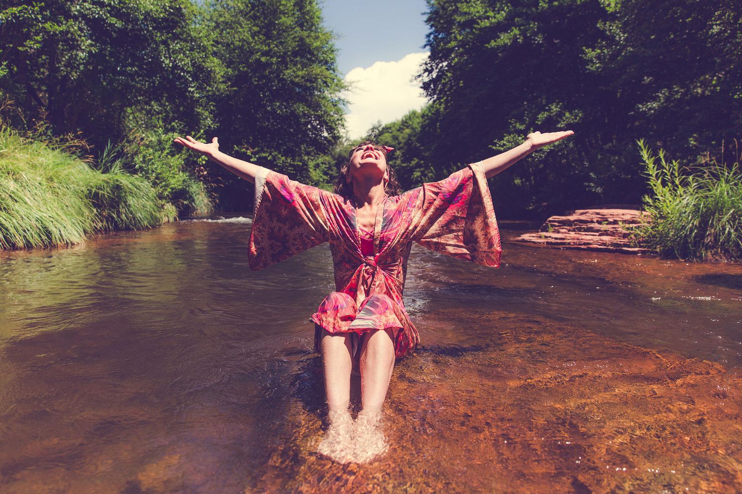 Woman in red floral dress standing in a shallow river with arms outstretched, surrounded by lush green trees.