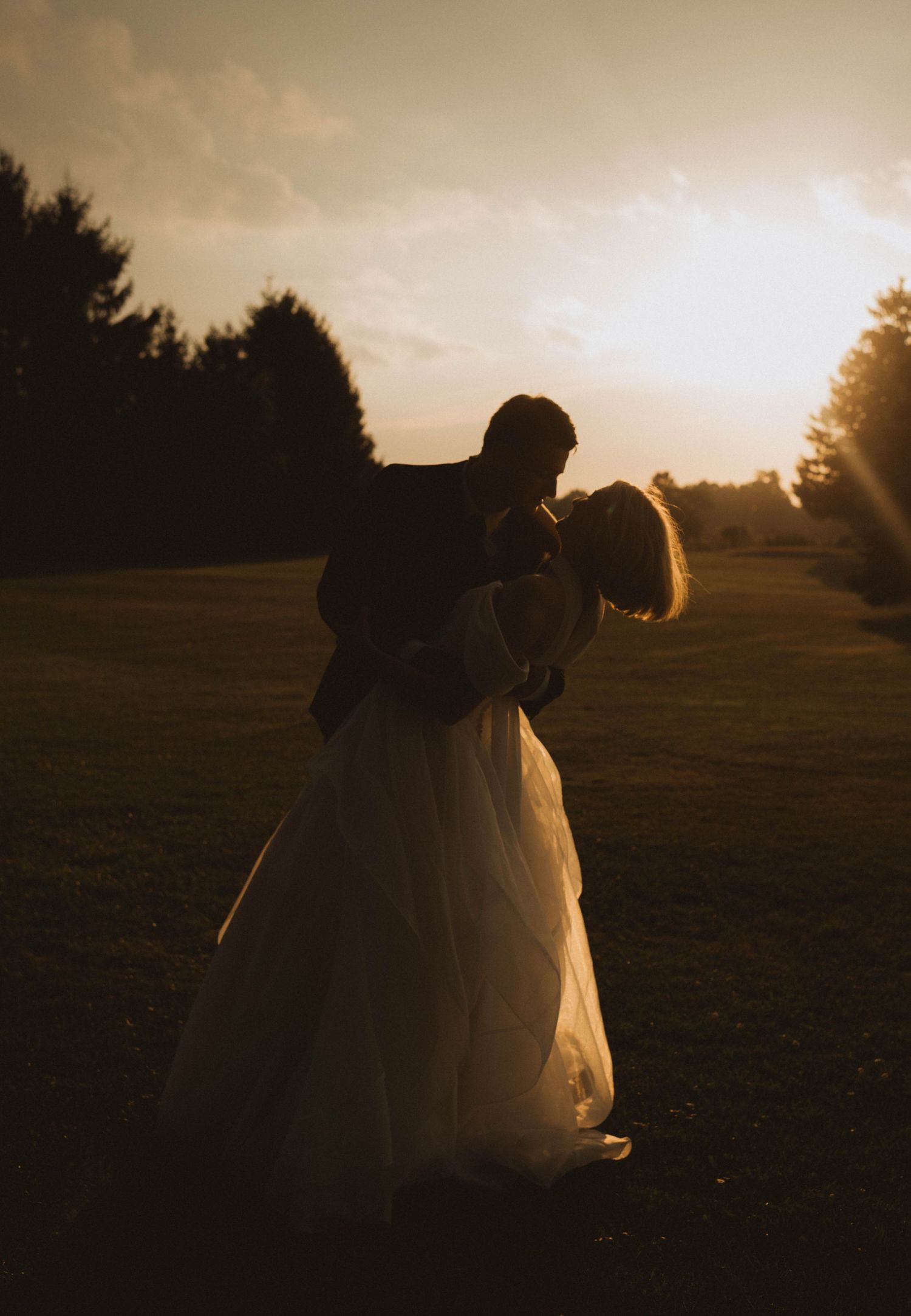 Silhouette of a bride and groom sharing a romantic kiss at golden sunset on a lush green lawn.