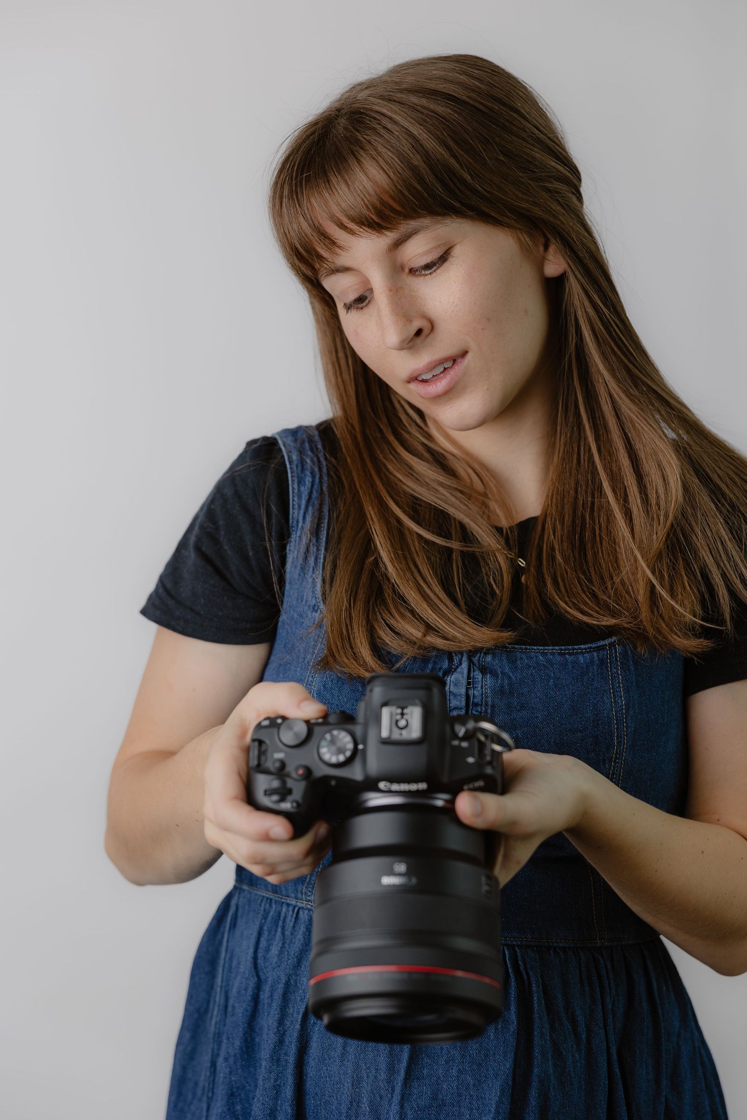 Young woman with brown hair holding a Canon DSLR camera, wearing a denim dress, reviewing photos on white background.