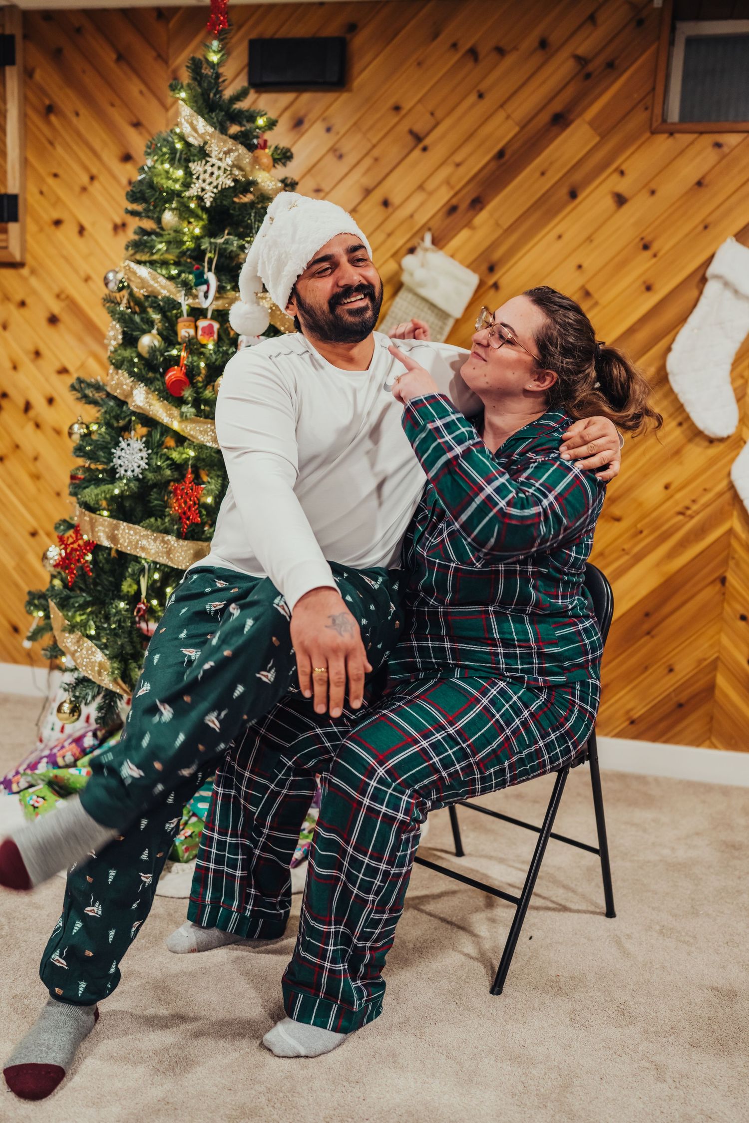 Couple in matching green plaid Christmas pajamas laughing together beside a decorated holiday tree with stockings on wall.