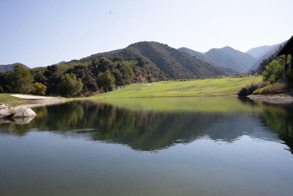 Tranquil mountain lake reflects green hills and forested slopes in a peaceful valley landscape.