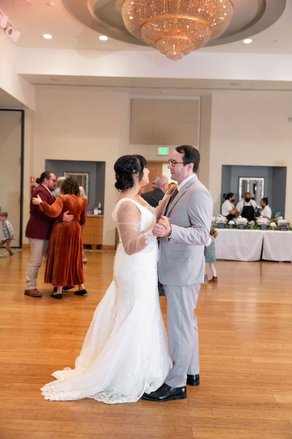 A couple shares their first dance as newlyweds in an elegant ballroom with a crystal chandelier and wood flooring.