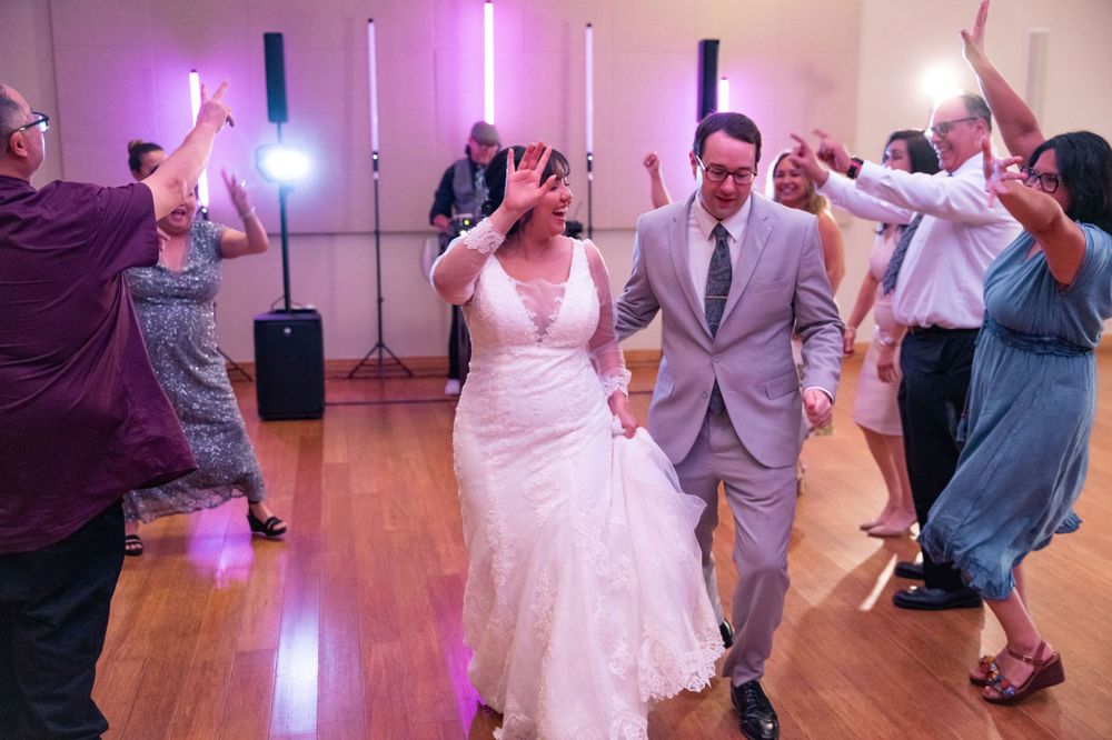 A wedding couple dances surrounded by guests at a reception with purple lighting and wood flooring.