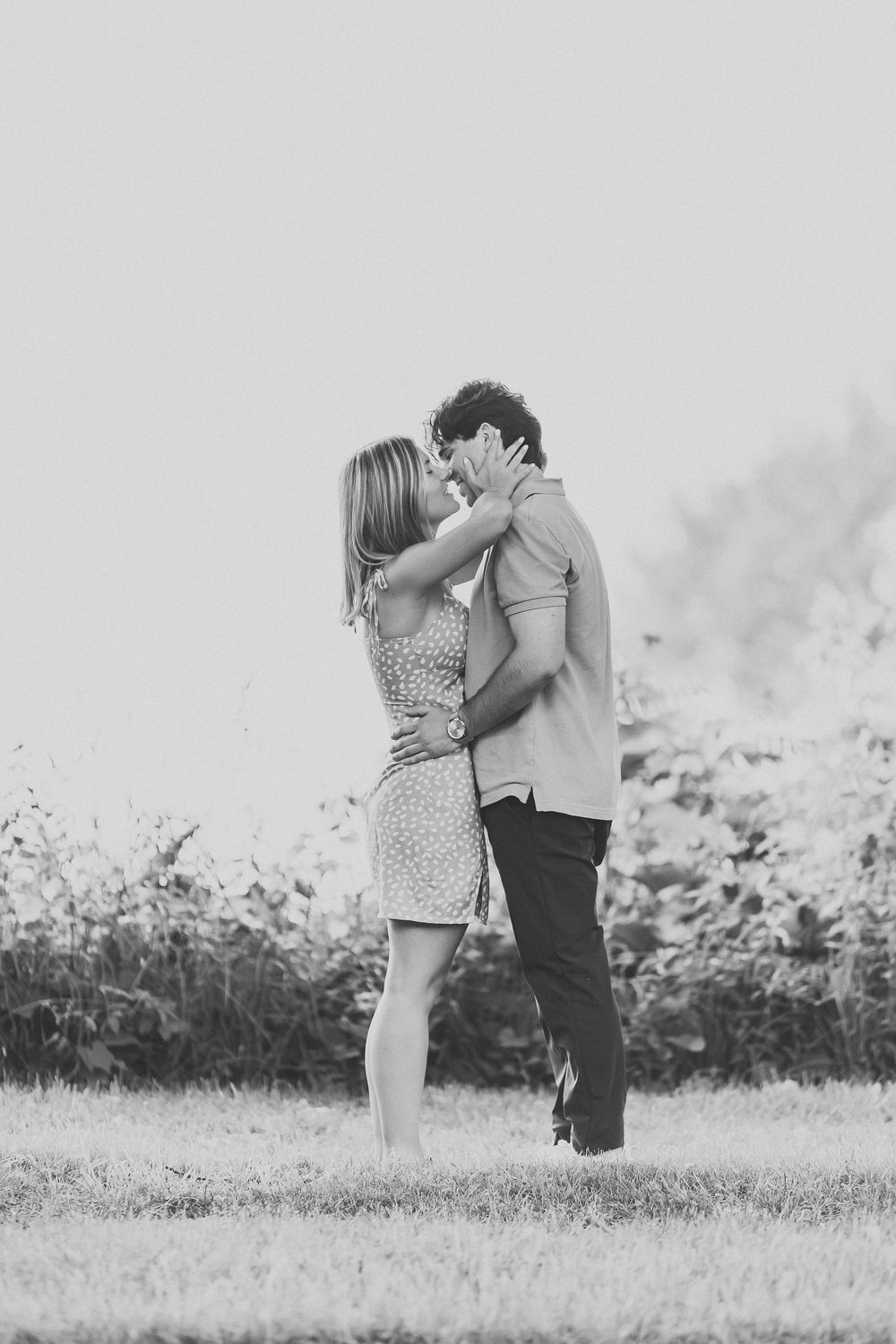 Black and white photo of a couple sharing a romantic kiss in a grassy field with wildflowers in the background.