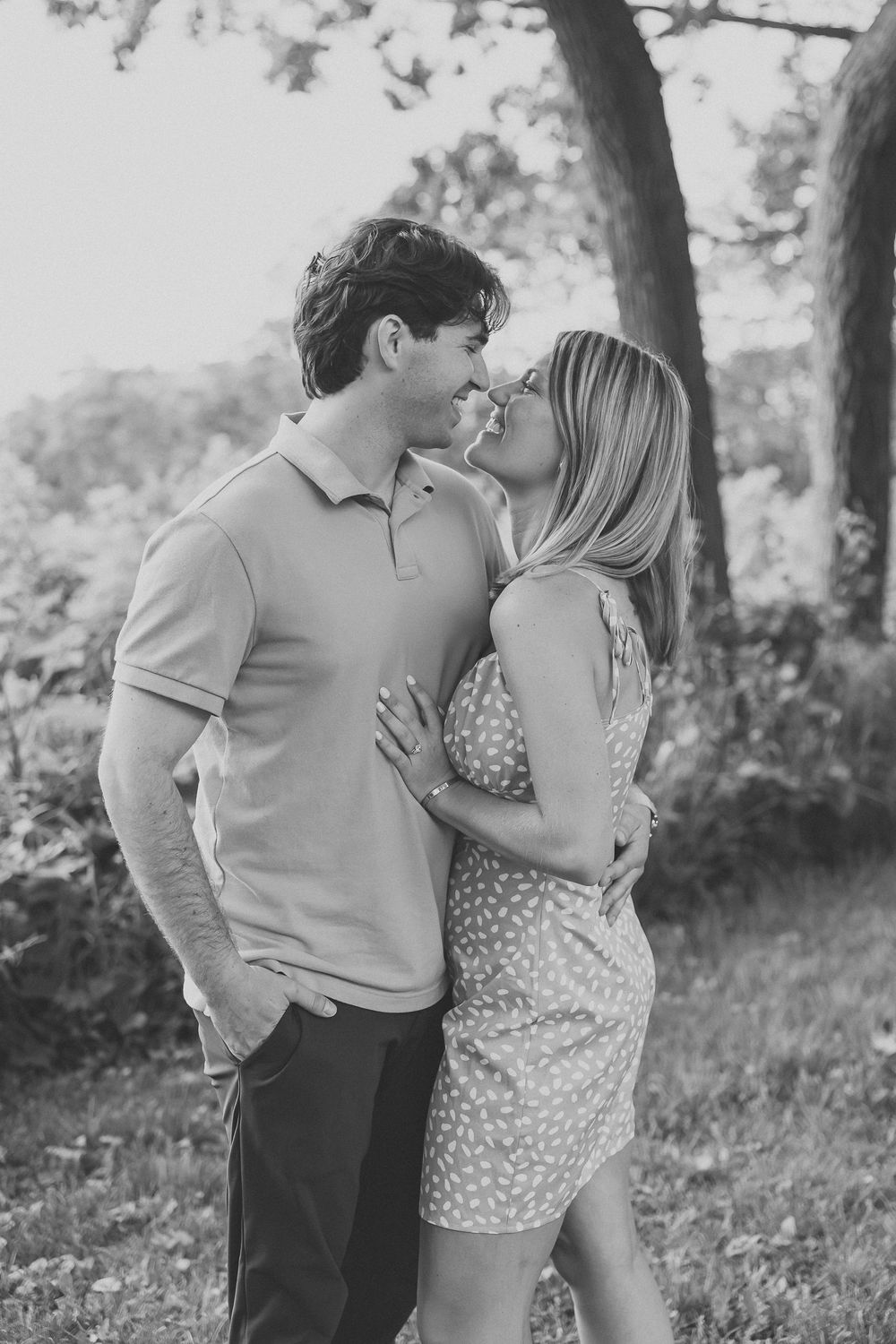 Black and white photo of a couple sharing a romantic kiss outdoors near a tree in a lush green setting.