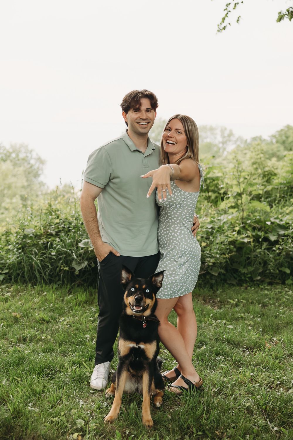 A smiling couple stands with their German Shepherd dog on a green lawn surrounded by lush foliage.