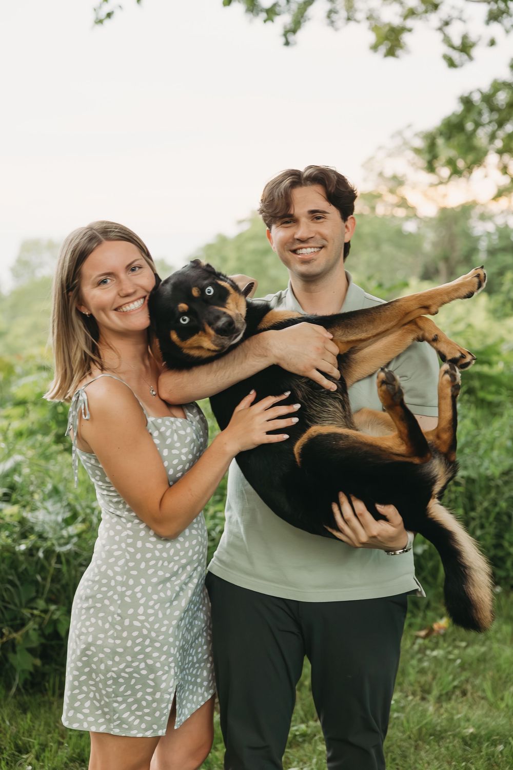 A smiling couple holds a playful black and tan dog outdoors surrounded by lush green foliage.