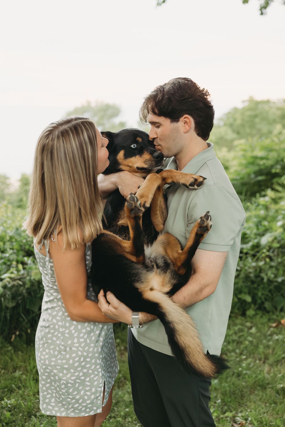 Couple holding a black and tan dog outdoors, sharing a kiss over their pet in a lush green garden setting.
