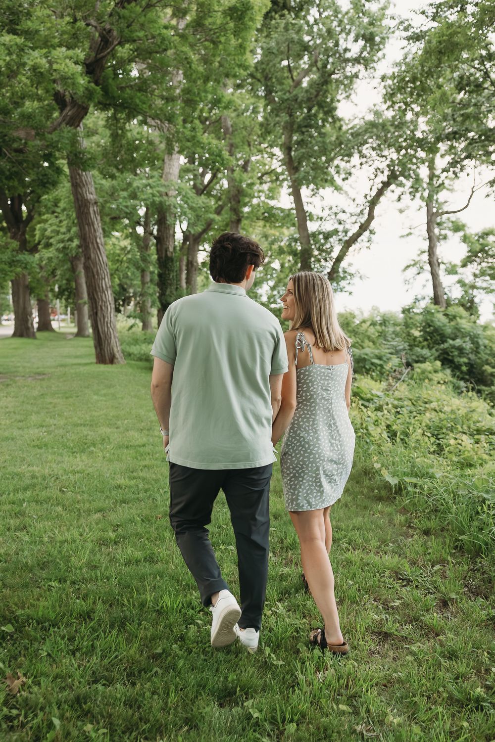 A couple walks hand in hand through a lush green park, the woman in a floral dress glancing back over her shoulder.